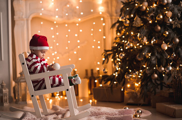 Baby 1 year old wearing santa claus suit sitting in rocking chair with Christmas tree and lights on background in room. Merry Christmas. Holiday season.