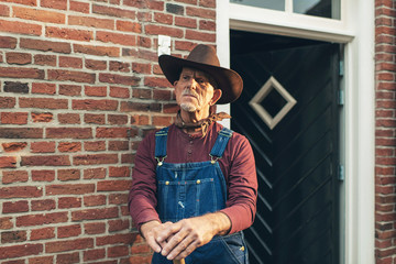 Senior farmer in dungarees with hat standing against wall of far