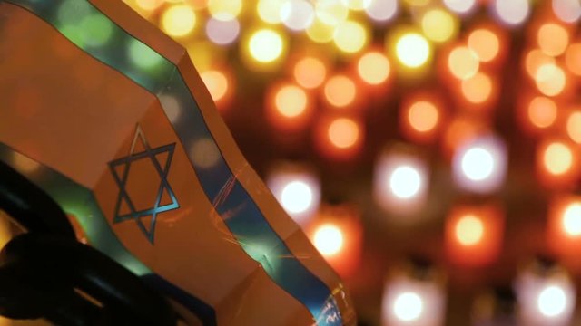 Israel flag mounted on a metal chain on a background of burning memorial candles. The concept of the Holocaust. A memorial ceremony dedicated to the Holocaust. Night Scene.