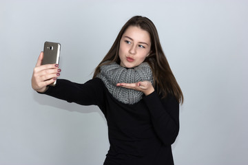 Portrait of a pretty woman in turtleneck sweater  with scarf she smiles and makes selfie  sending us a blowing kiss  standing over gray background and looking at camera
