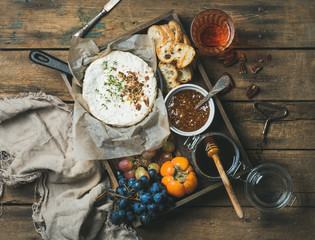 Cheese, fruit and wine set. Camembert in small pan, grapes, persimmon, fig jam, honey, baguette slices in wooden tray and glass of rose wine over rustic background, top view, horizontal composition