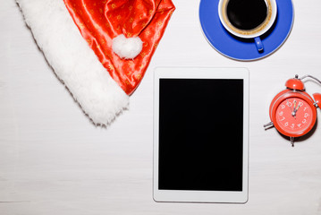 Santa Claus hat, tablet pc computer, coffee cup and alarm clock on white wooden table background