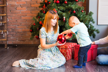 Happy mother and adorable baby holding bauble against domestic festive backdrop with Christmas tree