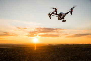 White drone with digital camera flying in sky over field on sunset
