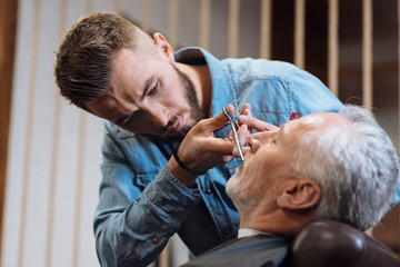 Handsome young barber cutting mustache of senior client with scissors