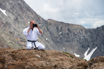 The man in a kimono sits in  lotus position on top of  mountain and taking pictures  the camera.