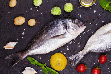 Raw fresh dorado fish with brussels sprouts, tomatoes, lemon, young potato, greens, bread, white wine bottle and olive oil on dark background. View from above, top studio shot