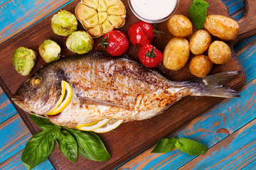 Broiled dorado fish with brussels sprouts, tomatoes, garlic, young potato and greens on cutting board. View from above, top studio shot