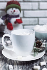Hot chocolate with marshmallows. Drink in a white ceramic cup with zephyr on wooden table. Decorative pine branch with the cone on plate, saucer. Snowman on background