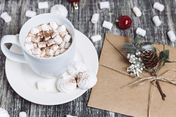 Obraz na płótnie Canvas Hot chocolate with marshmallows. Drink in a white ceramic cup with zephyr on wooden table. Envelop with decorative pine branch with the cone