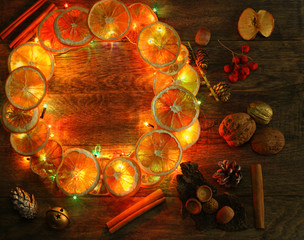 Christmas hand made craft template evening light background. Make traditional New Year`s door wreath components: tree cones, dried oranges, cinnamon, ribbon and colorful garland lights. Copyspace.