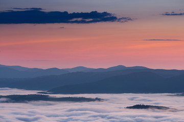 Summer weather phenomenon. Seasonal landscape with morning fog in valley. Clouds drenched valley below the level of the mountains. Sunrise over creeping clouds.