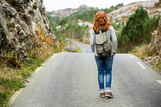 Woman traveler with a backpack on his shoulders walking along the road. The road through the rocks. Scandinavia, autumn, cloudy.