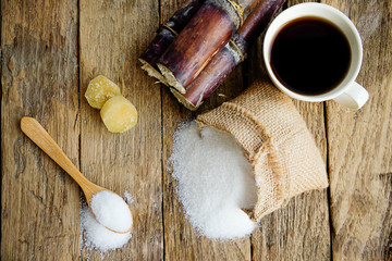 Fototapeta na wymiar Sugar in wooden spoon with sugar in burlap sack, sugarcane and white cup of coffee on wooden desk. Sugar crystals closeup. Top view and soft focus(selective focus).