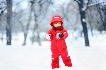 Portrait of little funny boy in red winter clothes having fun with snow during snowfall