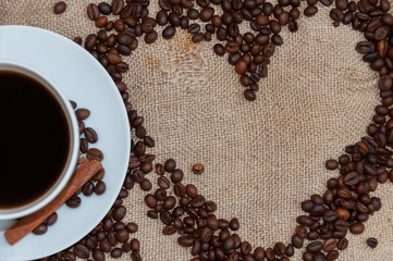 Naklejka premium Coffee beans in form of heart and white cup with cinnamon sticks on sackcloth
