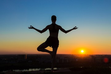 Fototapeta na wymiar Silhouette of sporty woman practicing yoga in the park at sunset - half lotus tree pose. Sunset light, golden hour. Freedom, health and yoga concept