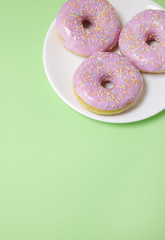 A plate of ring donuts with pastel pink frosting and sprinkles on a soft green background with empty space below