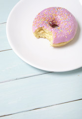 A ring doughnut with pastel pink frosting and sprinkles on a painted wooden table background, with a bite missing