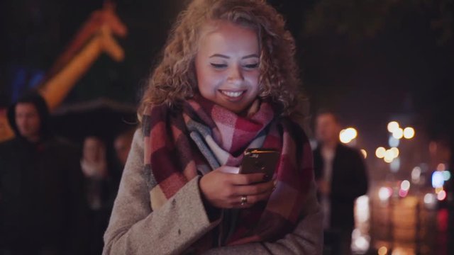 Curly blonde girl walking down the street and texting a message, smiles and muffles up in a warm scarf. People passing by under the umbrellas. Happy loneliness. City lights on the background.
