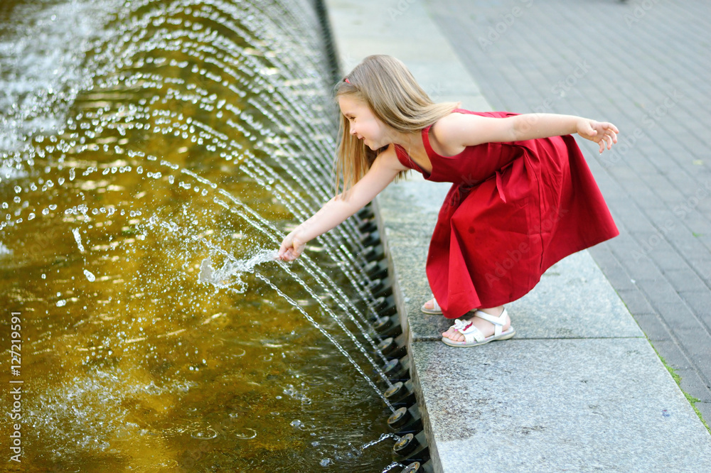 Wall mural Cute little girl playing with a city fountain on hot and sunny summer day