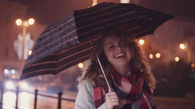 Sweet smile of cute blondie walking down the street. Outside shootage, passing by cars and city lights on the background. Rainy atmosphere. Slow motion.