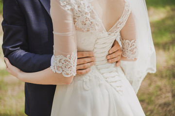 Happy wedding couple standing in park together. Close up of grooms hands on waist of beautiful bride. Color horizontal photo.
