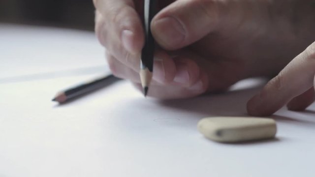 Close up of a Caucasian male hand drawing and erasing with a black pencil on white paper