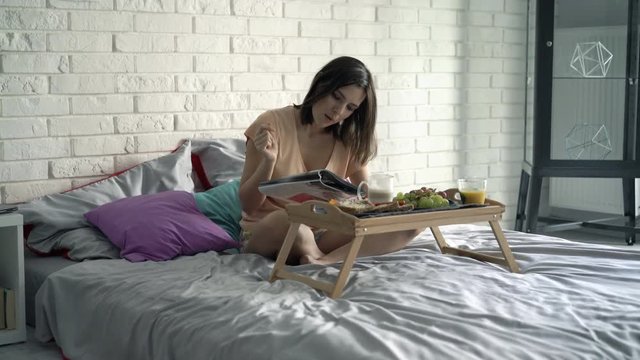 Young woman reading magazine during breakfast sitting on bed at home

