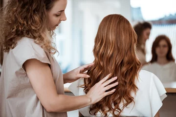 Tuinposter Female hairdresser making hairstyle to redhead girl in beauty salon. © Cookie Studio
