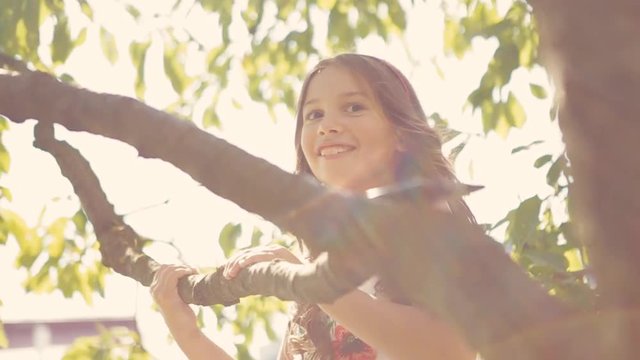 European teenage girl in white dress, with curly hair climbs a tree in a bright sunshine. Moments of happiness. Happy childhood. Outdoors activities. Children portrait.
