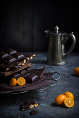 various sorts of dark chocolate bars, kumquats and a small silver coffee jug on a table against a dark background