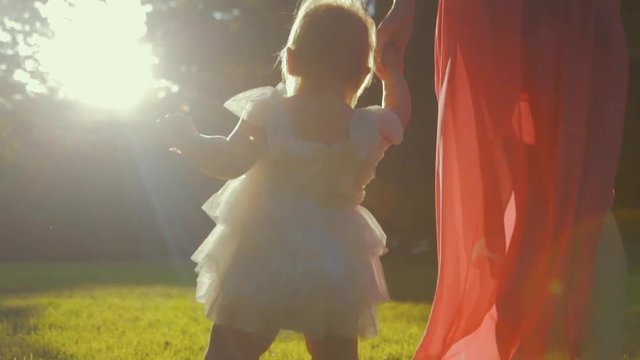 Baby in a puffy white dress makes first steps holding mother hand in a bright sunlight. The setting sun plays in downy hair of baby-girl. Elegant mother walking with her baby-girl in the city park.