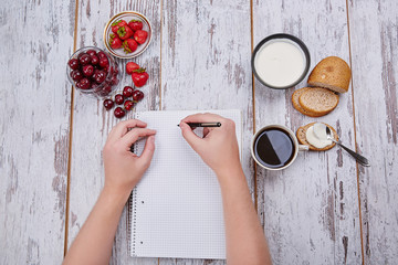 Cup coffee, metal spoon, strawberry, cherry, sour cream and silced bread and notepad on rustic table with hands