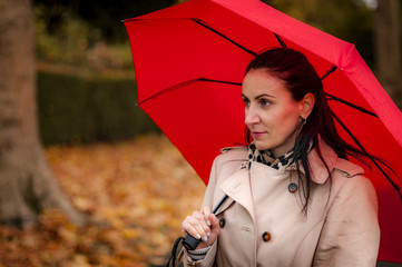 Autumn setting with a mature woman standing outdoors in a park next to a tree in the fall, wearing a trench coat holding and a red umbrella not looking at the camera, admiring the fall foliage