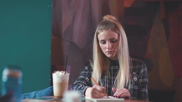 Portrait of a young girl in the cafe: woman drinking coffee and making sketches in a notebook. Young blonde woman sitting at a table in a coffee shop and writes something in a notebook.