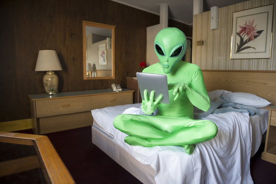 Green alien sitting using futuristic tablet computer on the bed in an old-fashioned room