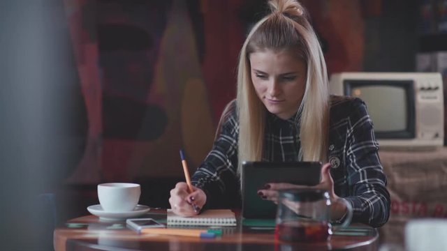 Modern girl in a cafe drinking tea, uses the tablet and writes in notebook. Beautiful girl in the coffee shop working or studying. Remote work. Stylish woman with a tab and notebook in a coffee shop