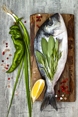 Fresh uncooked dorado or sea bream fish with lemon, aromatic herbs, vegetables and spices on rustic wooden board over grey stone background. Top view