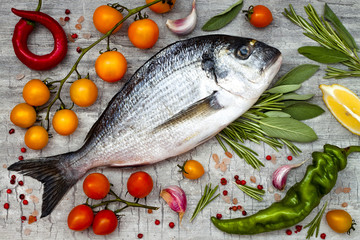Fresh uncooked dorado or sea bream fish with lemon, aromatic herbs, vegetables and spices over grey stone background. Top view