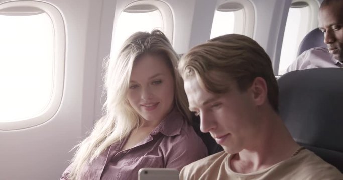 Attractive young couple watch online content on a smart phone as they fly in main cabin of commercial jet airliner. Medium close up from front angle, recorded hand-held at 60fps