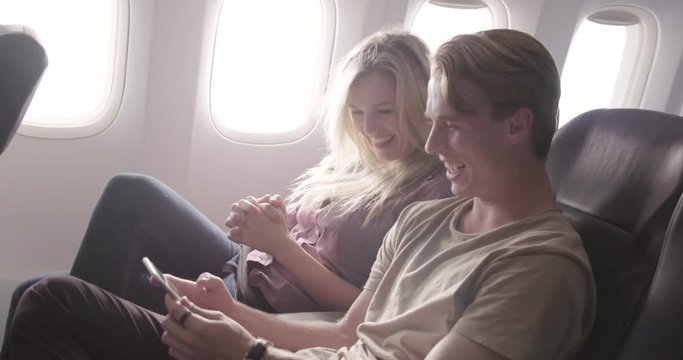 Attractive young couple enjoy watching online content on a smart phone as they fly in main cabin of commercial jet airliner. Medium shot from side angle, recorded hand-held at 60fps