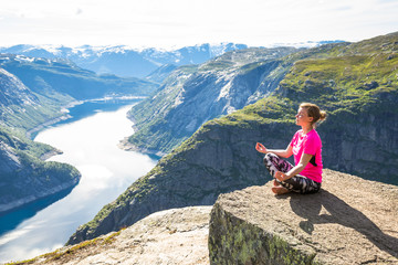 Young woman doing yoga on Trolltunga. Happy girl enjoy beautiful lake and good weather in Norway.