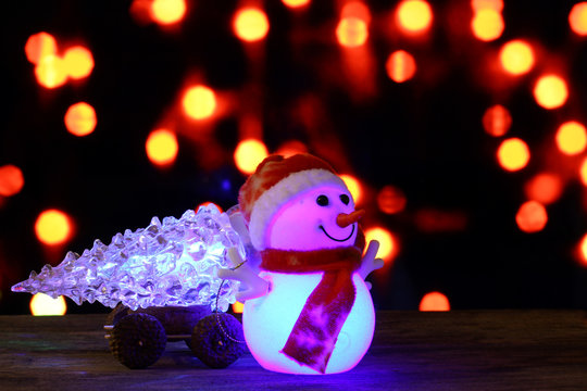 Dark evening Christmas and New Year`s card or poster with Snowman drags a truck glowing glass tree on bokeh background. Big copyspace place for text, logo, sale price and item description.