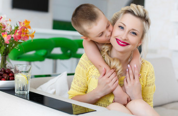 Home portrait of a happy mother and her son,6 years old,sitting together on a large bright kitchen,a woman wearing a yellow blouse,blonde with blue eyes,son hugging mother standing behind back