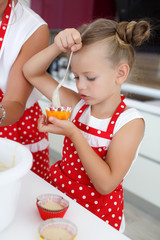 Young woman,brunette,dressed in a white shirt and a red apron with white polka dots,with my daughter 5 years in white tank top and a red apron with white polka dots baking cupcakes in the kitchen