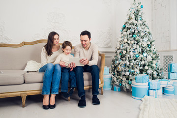 Obraz premium Cheerful family sitting in the living room having fun with the digital tablet that Santa Claus brought her, behind the decorated christmas tree