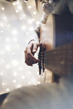 Hipster girl playing guitar in a home atmosphere, person studying on musical instrument on glow bokeh Christmas illimination, hands using guitar in holiday on relax glitter xmas decoration, blur
