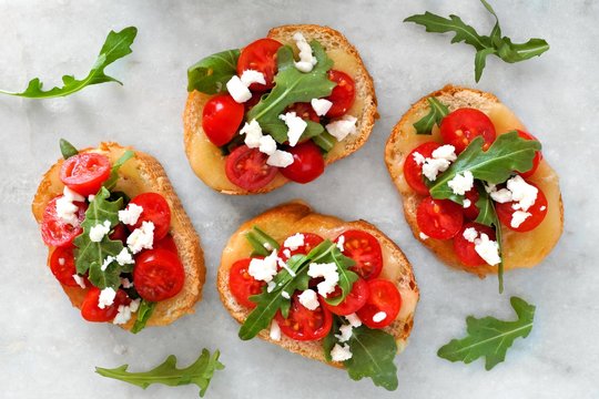 Crostini appetizers with cherry tomatoes, arugula, and cheese, above view on white marble