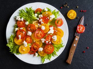 Colorful salad, fresh green leaves and sliced red and yellow cherry tomatoes, white plate, knife, black stone background, top view.